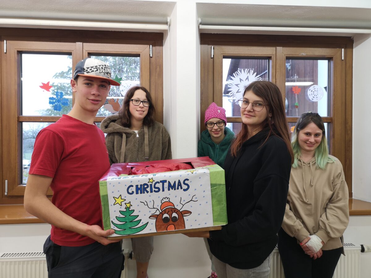 fünf junge Erwachsene lächeln in die Kamera, zwei halten ein großes Weihnachtspaket